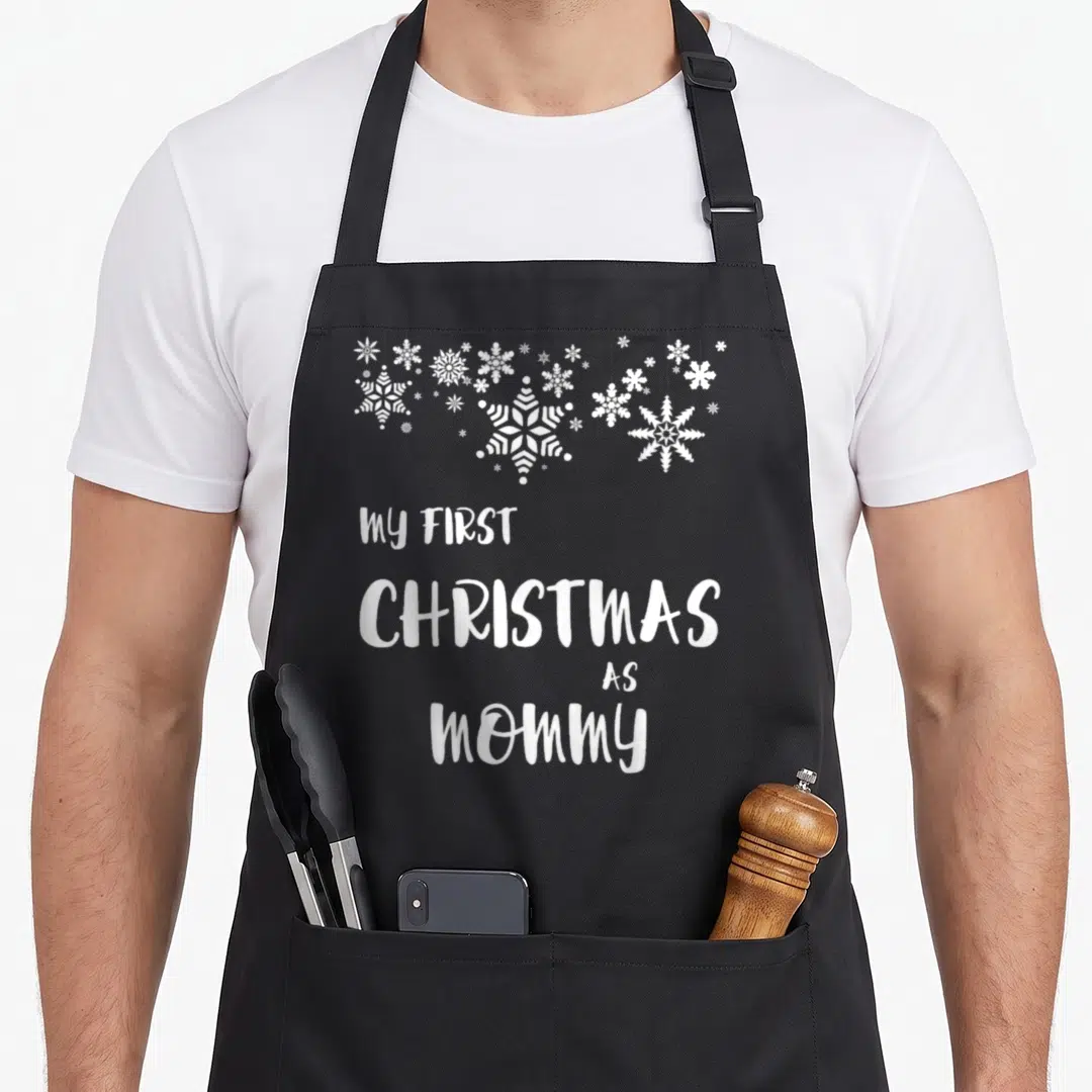 Funny christmas aprons - White snowflake cluster above stacked text reading My First Christmas As Mommy — shown on a black cotton canvas apron against a white background