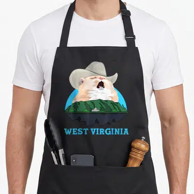 West cat apron - Singing beige cat in cowboy hat behind green mountains with West Virginia text — shown on a black cotton canvas apron against a white background