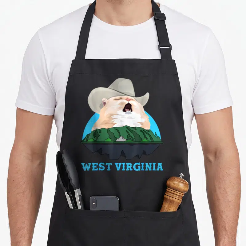 West cat apron - Singing beige cat in cowboy hat behind green mountains with West Virginia text — shown on a black cotton canvas apron against a white background