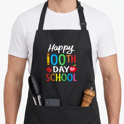 Baseball teacher apron - Stacked text Happy 100th Day of School with crayon notebook and apple in white blue green red — shown on a black cotton canvas apron against a white background
