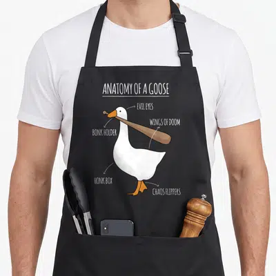 Anatomy goose apron - Cartoon white goose holding a bat with labels Evil Eyes and Chaos Flipper — shown on a black cotton canvas apron against a white background