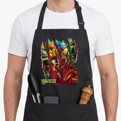 Shredder teenage cooking aprons - Four turtles and Shredder head through red tears with TMNT logo text — shown on a black cotton canvas apron against a white background