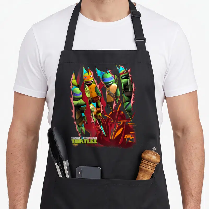 Shredder teenage cooking aprons - Four turtles and Shredder head through red tears with TMNT logo text — shown on a black cotton canvas apron against a white background