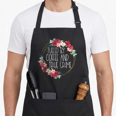 Murderino coffee apron - Floral wreath in red pink and teal surrounding stacked white text FUELED BY COFFEE AND TRUE — shown on a black cotton canvas apron against a white background
