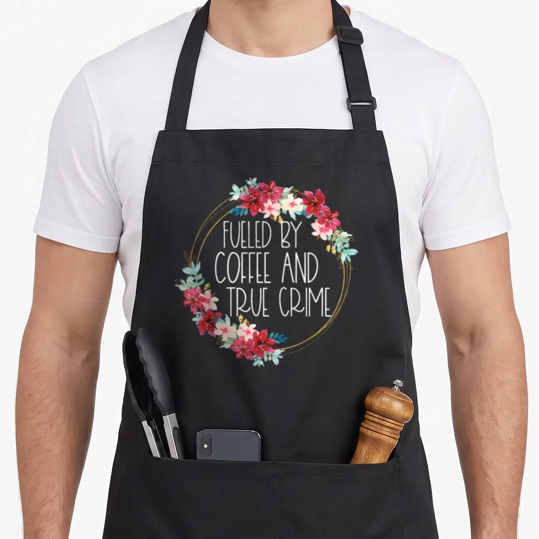 Murderino coffee apron - Floral wreath in red pink and teal surrounding stacked white text FUELED BY COFFEE AND TRUE — shown on a black cotton canvas apron against a white background