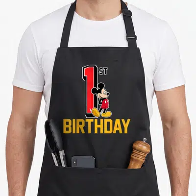Funny 1st birthday apron - Mickey Mouse leaning against red number 1 with yellow BIRTHDAY text below — shown on a black cotton canvas apron against a white background