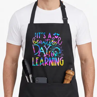 Beautiful tie-dye apron - Stacked tie-dye text reading It's a beautiful DAY FOR LEARNING with hearts, rainbow — shown on a black cotton canvas apron against a white background
