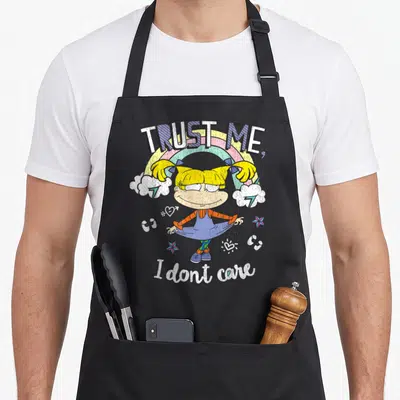 Angelica rainbow apron - Cartoon girl under rainbow with TRUST ME I don't care text and scattered stars — shown on a black cotton canvas apron against a white background