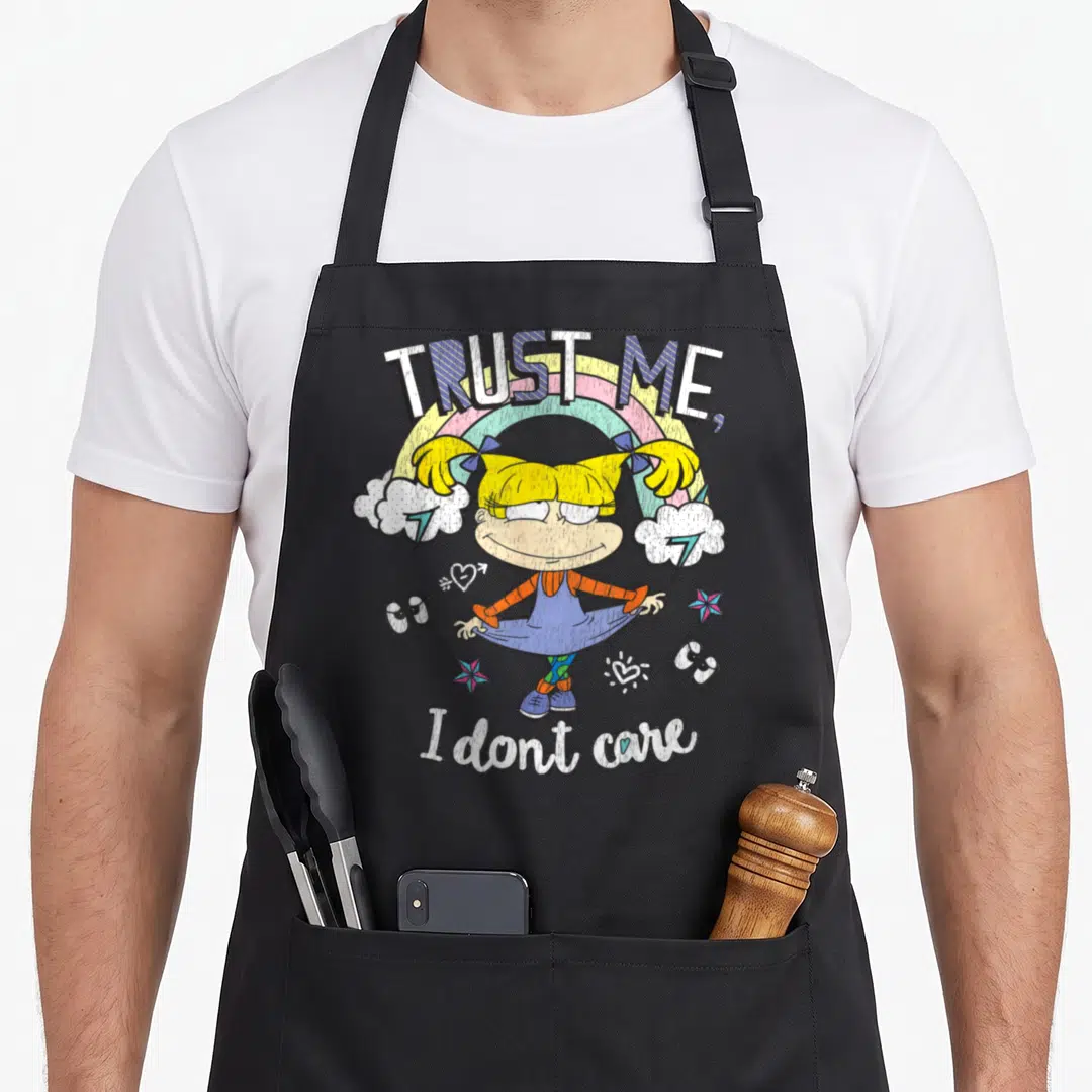 Angelica rainbow apron - Cartoon girl under rainbow with TRUST ME I don't care text and scattered stars — shown on a black cotton canvas apron against a white background