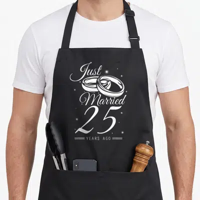 25th anniversary wedding apron - White Just Married 25 Years Ago text with two interlocking rings and sparkles — shown on a black cotton canvas apron against a white background