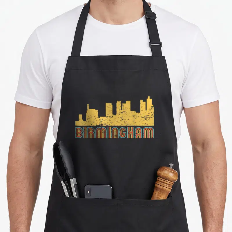Skyline alabama apron - Yellow skyline silhouette above BIRMINGHAM text in teal and orange retro font — shown on a black cotton canvas apron against a white background
