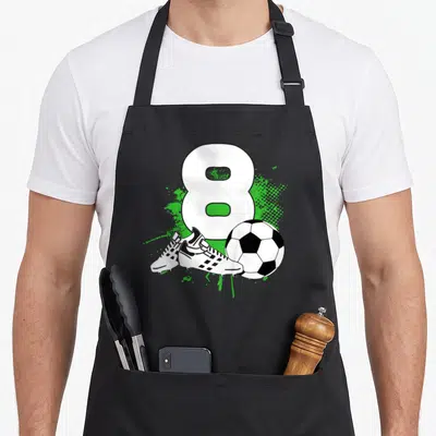 8th soccer birthday apron - White number 8 with soccer ball and cleats over green splatter — shown on a black cotton canvas apron against a white background