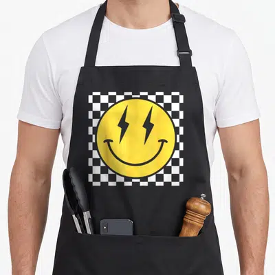 Distressed face on apron - Yellow smiley face with grey lightning bolt eyes on white and grey checkerboard — shown on a black cotton canvas apron against a white background