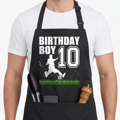 Theme 10th birthday apron - BIRTHDAY BOY 10 text with white soccer player silhouette kicking ball over grass — shown on a black cotton canvas apron against a white background
