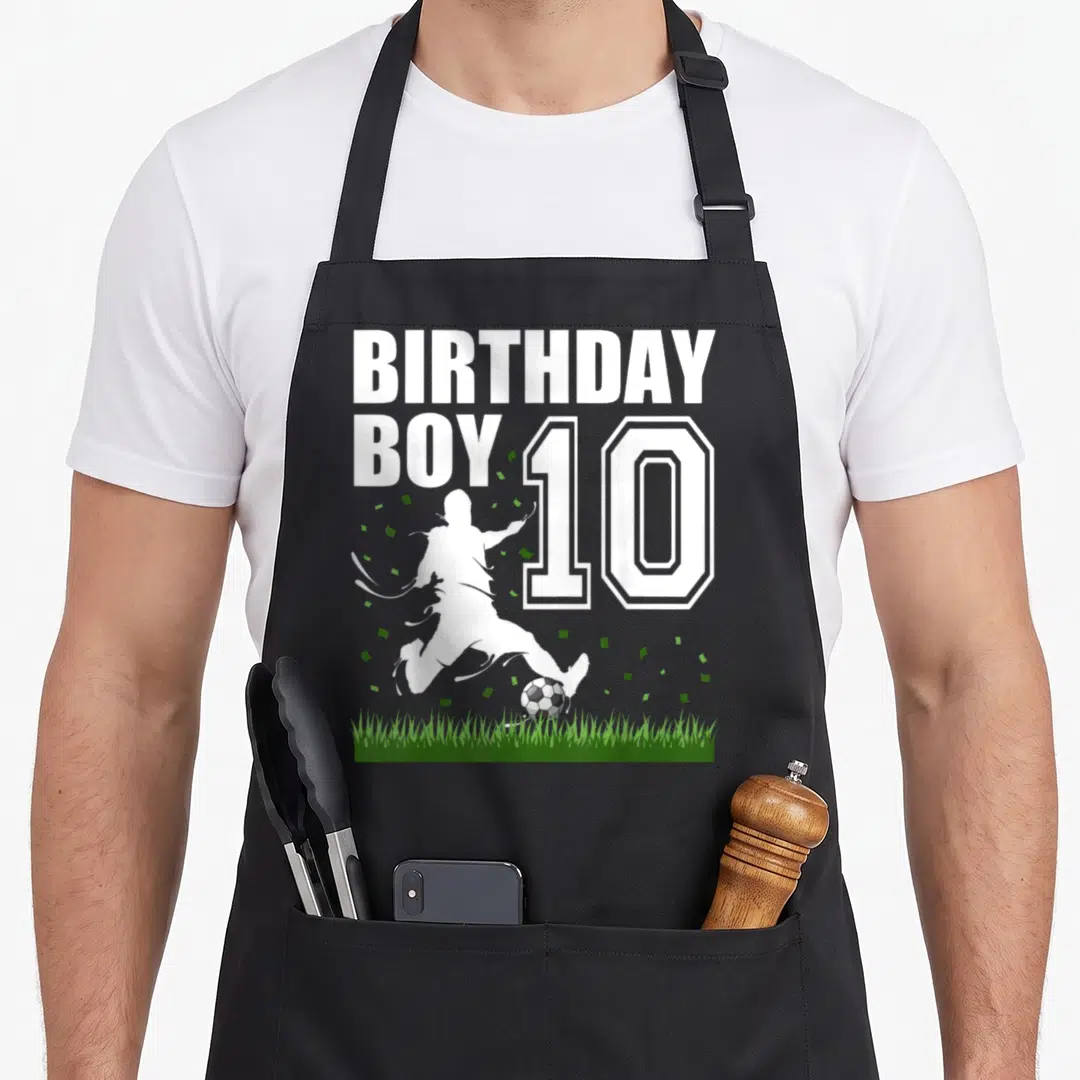 Theme 10th birthday apron - BIRTHDAY BOY 10 text with white soccer player silhouette kicking ball over grass — shown on a black cotton canvas apron against a white background
