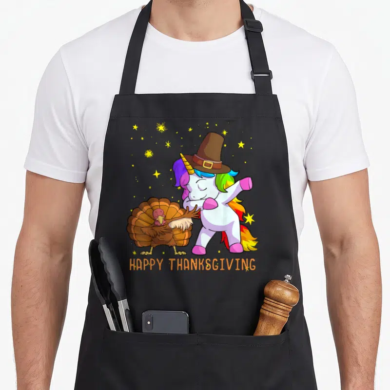 Unicorn pilgrim apron - Dancing unicorn in brown pilgrim hat next to turkey with yellow stars and orange text — shown on a black cotton canvas apron against a white background