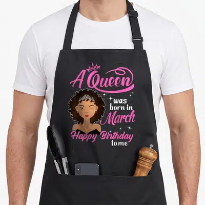 Cute march baking queen apron - Cartoon woman winking with A Queen was born in March text in pink and white — shown on a black cotton canvas apron against a white background