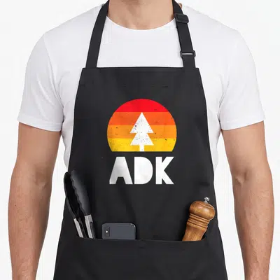 Funny adk adirondacks apron - White tree silhouette in red orange yellow striped semi circle with ADK text below — shown on a black cotton canvas apron against a white background
