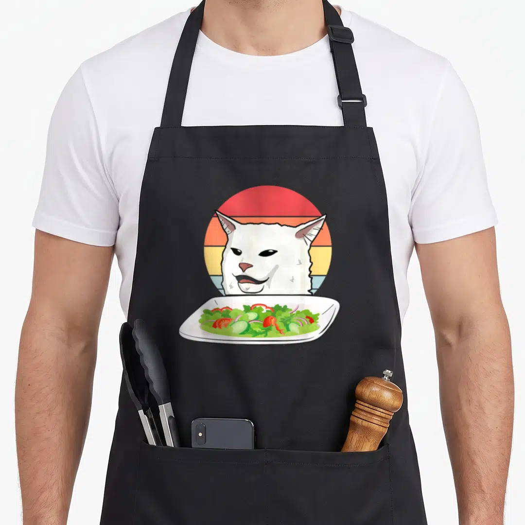 Angry cat apron - White cartoon cat head over salad plate in front of striped retro sun — shown on a black cotton canvas apron against a white background