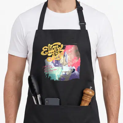 Elton colorful apron - Space shuttle, Elton John figure, yellow script text, pink starry background — shown on a black cotton canvas apron against a white background
