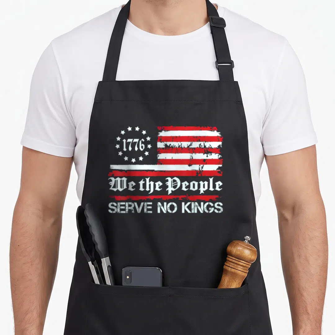 King to serve man apron - Distressed red white USA flag with 1776 stars and We the People Serve No Kings text — shown on a black cotton canvas apron against a white background