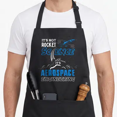 Funny rocket science apron - Blue rocket illustration with stacked text reading It's Not Rocket Science Aerospace — shown on a black cotton canvas apron against a white background