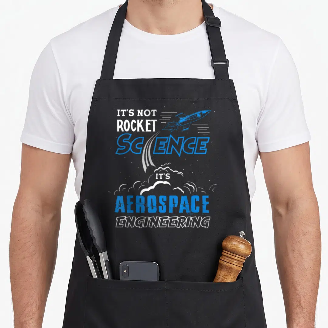 Funny rocket science apron - Blue rocket illustration with stacked text reading It's Not Rocket Science Aerospace — shown on a black cotton canvas apron against a white background