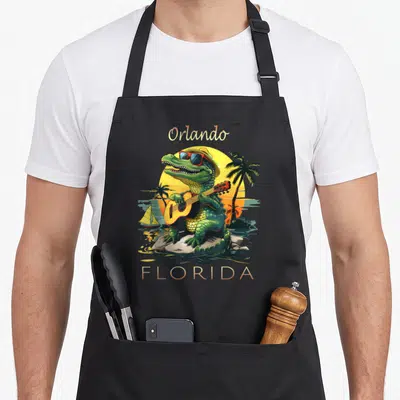 Vacation alligator florida apron - Cartoon green alligator playing guitar on rocks with Orlando FLORIDA text and sunset — shown on a black cotton canvas apron against a white background