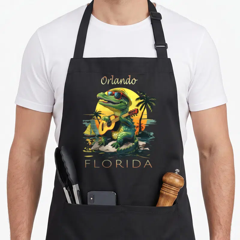 Vacation alligator florida apron - Cartoon green alligator playing guitar on rocks with Orlando FLORIDA text and sunset — shown on a black cotton canvas apron against a white background
