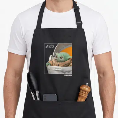 Snack novelty apron - Grogu in white pod with SNACK? text in white frame — shown on a black cotton canvas apron against a white background