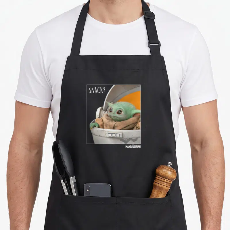 Snack novelty apron - Grogu in white pod with SNACK? text in white frame — shown on a black cotton canvas apron against a white background