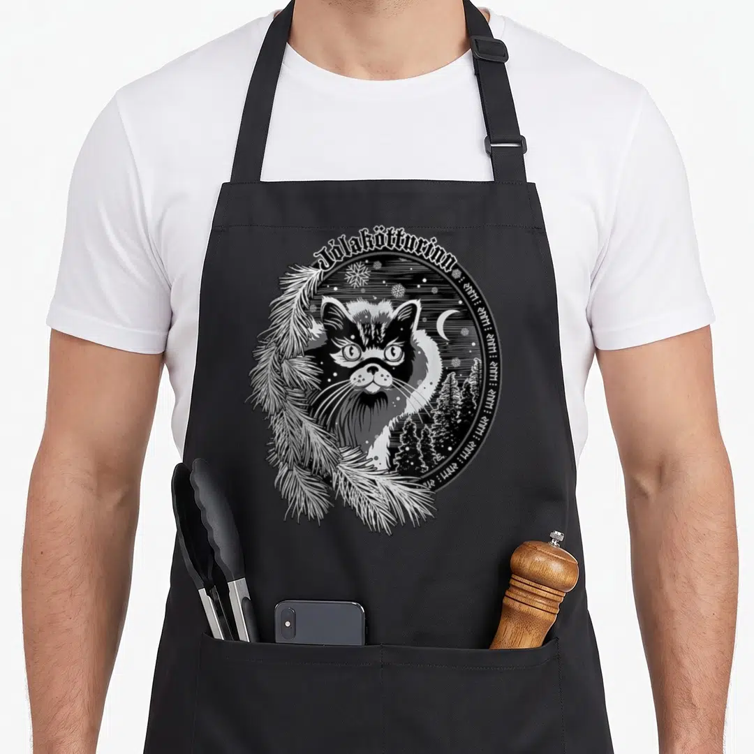 Yule christmas cat apron - Jólakötturinn cat face circle with pine branches, moon, stars, and runic text in line art — shown on a black cotton canvas apron against a white background