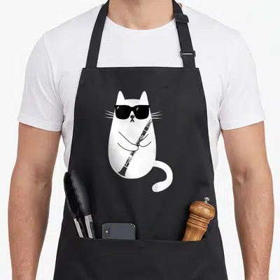 Musician cat wearing apron - White cartoon cat with black sunglasses holding a black clarinet diagonally — shown on a black cotton canvas apron against a white background