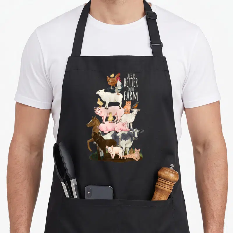 Better farm aprons - Watercolor stack of farm animals next to white LIFE IS BETTER ON THE FARM text on black fabric — shown on a black cotton canvas apron against a white background