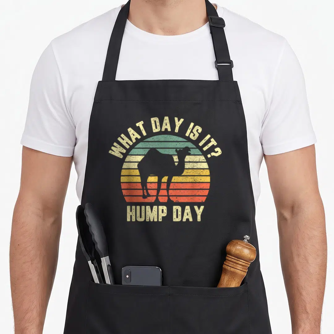 Hump camel apron - Retro striped sun with camel silhouette and What Day Is It Hump Day text — shown on a black cotton canvas apron against a white background