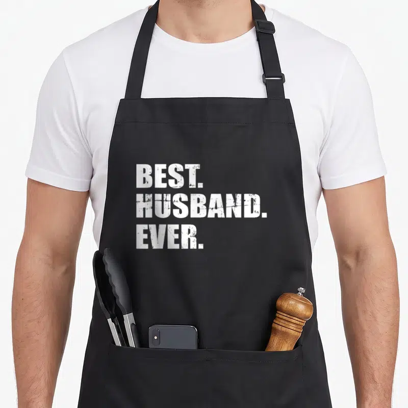 Spouse apron - White distressed BEST HUSBAND EVER text stacked on black fabric — shown on a black cotton canvas apron against a white background