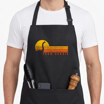 Loch monster lover apron - Retro Team Nessie text below grey silhouette overlapping yellow sun and orange stripes — shown on a black cotton canvas apron against a white background