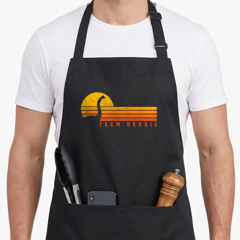 Loch monster lover apron - Retro Team Nessie text below grey silhouette overlapping yellow sun and orange stripes — shown on a black cotton canvas apron against a white background