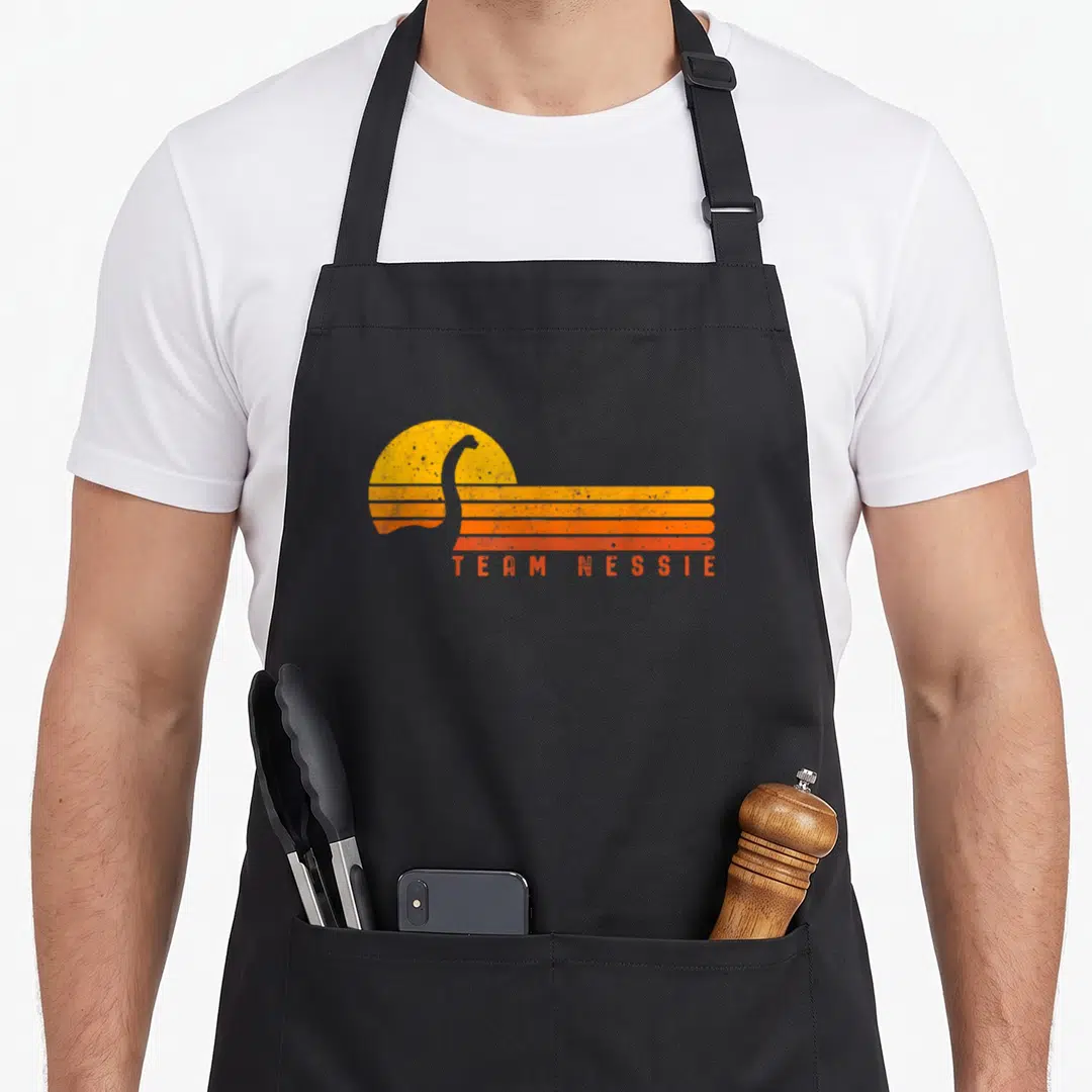 Loch monster lover apron - Retro Team Nessie text below grey silhouette overlapping yellow sun and orange stripes — shown on a black cotton canvas apron against a white background