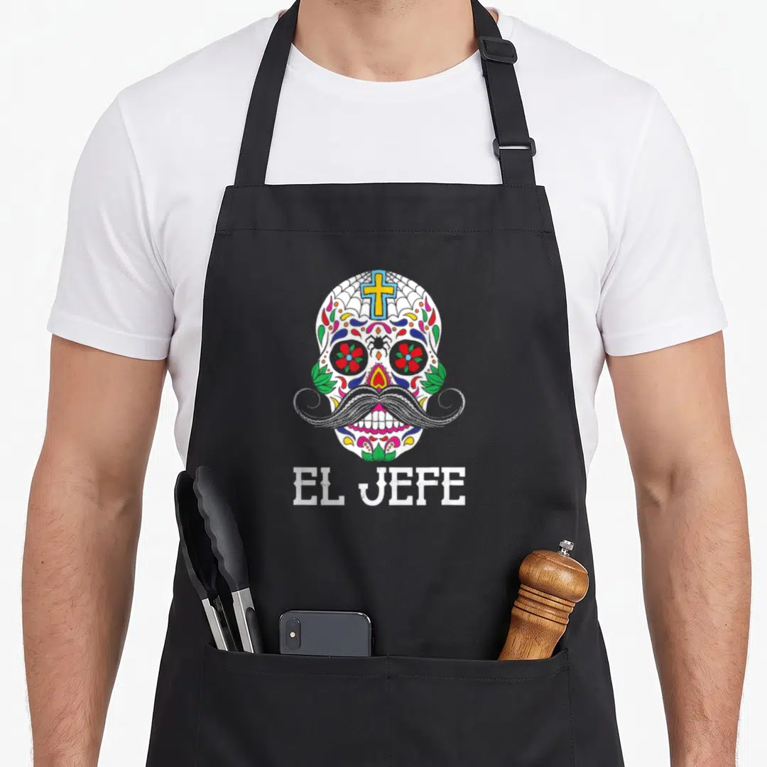 Soy day of the dead apron - Mustachioed sugar skull with EL JEFE text in white red green yellow blue cartoon style — shown on a black cotton canvas apron against a white background