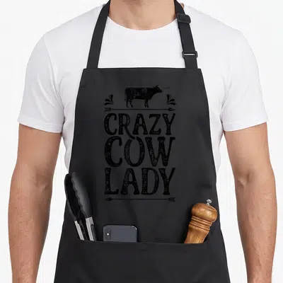 Farm cow print aprons - Black distressed cow silhouette above stacked CRAZY COW LADY text with arrows and flourishes — shown on a black cotton canvas apron against a white background