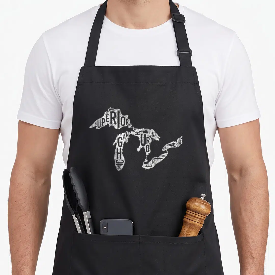 Funny lake distressed apron - White text forming Great Lakes map shape spelling Superior Michigan Huron Erie Ontario — shown on a black cotton canvas apron against a white background