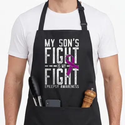 Son epilepsy awareness apron - Distressed white text My Son's Fight Is My Fight with purple epilepsy awareness ribbon — shown on a black cotton canvas apron against a white background