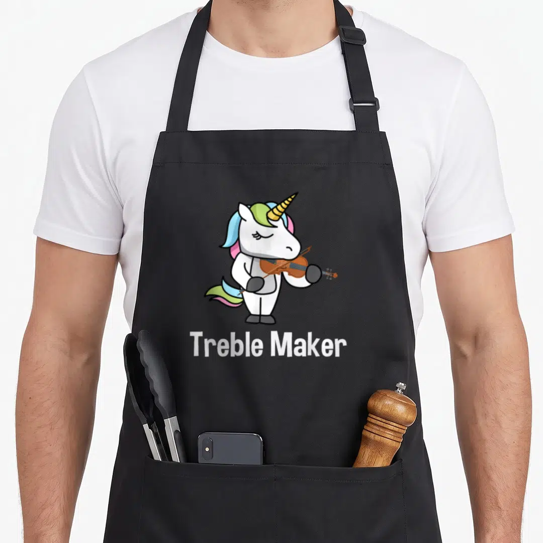 Violinist maker apron - Cartoon white unicorn playing brown violin with rainbow mane and 'Treble Maker' text below — shown on a black cotton canvas apron against a white background