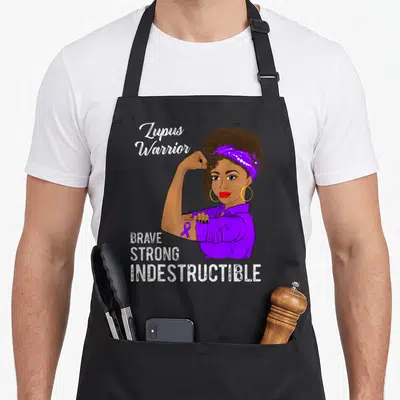 Indestructible lupus awareness apron - Cartoon woman flexing arm with purple ribbon and text Lupus Warrior Brave Strong — shown on a black cotton canvas apron against a white background