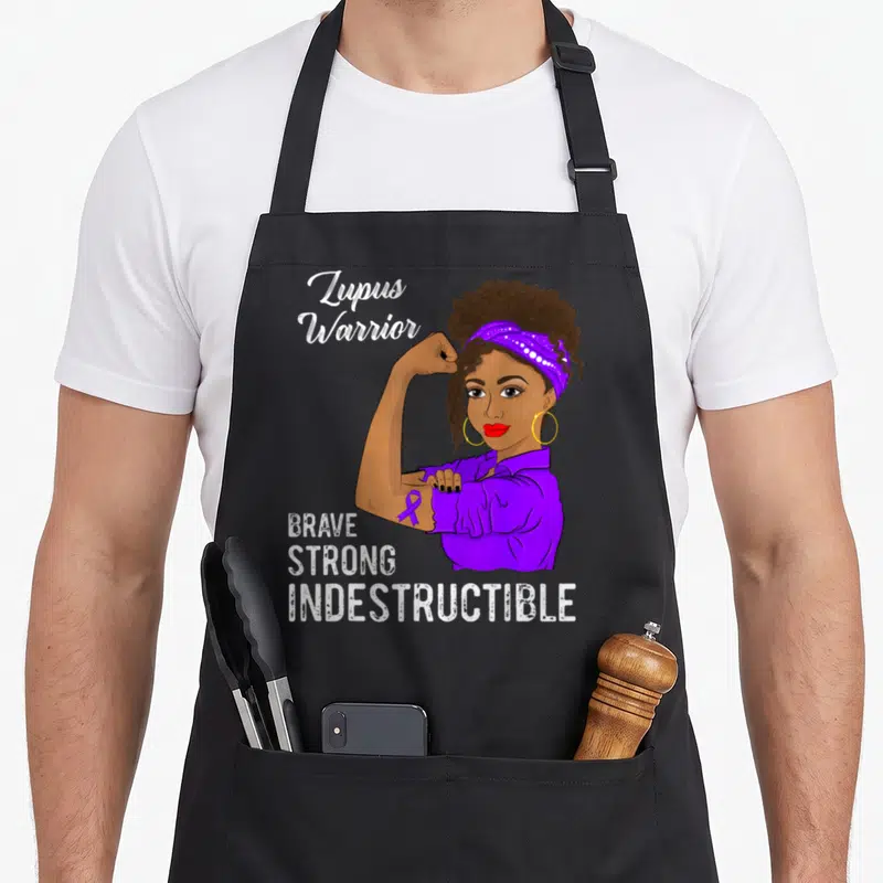 Indestructible lupus awareness apron - Cartoon woman flexing arm with purple ribbon and text Lupus Warrior Brave Strong — shown on a black cotton canvas apron against a white background