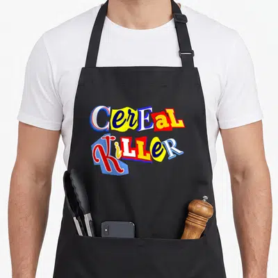Killer food aprons - Stacked Cereal Killer text in blue yellow red and white block letters — shown on a black cotton canvas apron against a white background