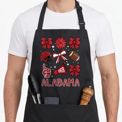 Cute youth alabama apron - Cheer bows, football helmet, pom, megaphone, foam finger, and ALABAMA text in red white — shown on a black cotton canvas apron against a white background