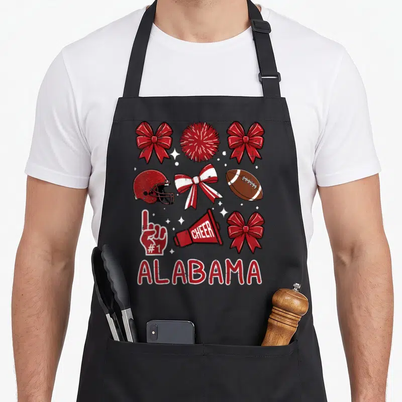 Cute youth alabama apron - Cheer bows, football helmet, pom, megaphone, foam finger, and ALABAMA text in red white — shown on a black cotton canvas apron against a white background