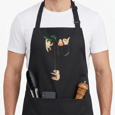 Family sloth apron - Four brown cartoon sloths hanging from green branches and a vine beside a grey pocket containing — shown on a black cotton canvas apron against a white background